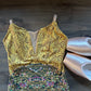 A sleeveless leotard or dance top laid flat on a dark wood floor, with a metallic gold upper section featuring swirling patterns and a multicolored sequined floral lower section. Two pale pink satin ballet slippers are placed beside it.