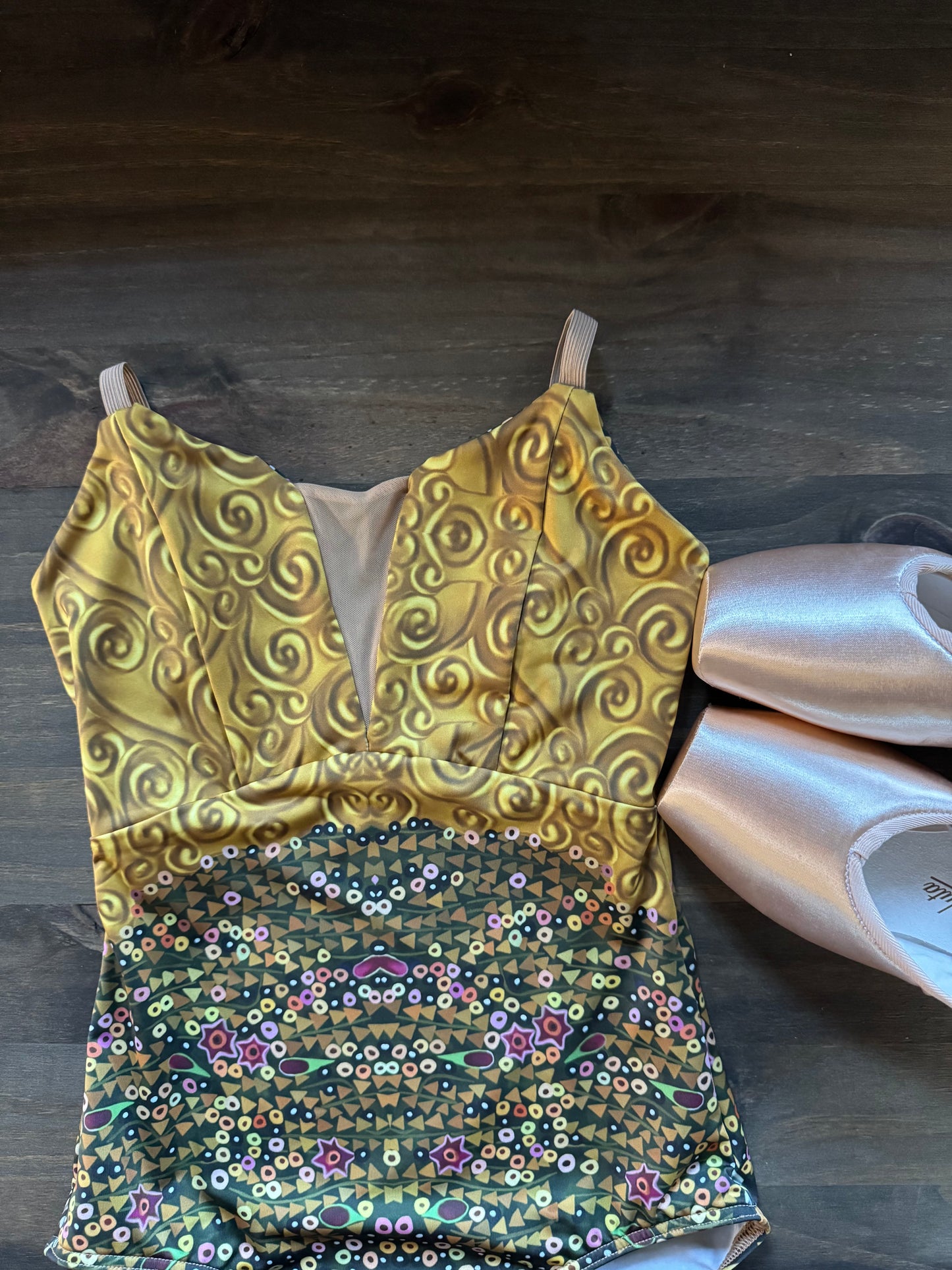 A sleeveless leotard or dance top laid flat on a dark wood floor, with a metallic gold upper section featuring swirling patterns and a multicolored sequined floral lower section. Two pale pink satin ballet slippers are placed beside it.