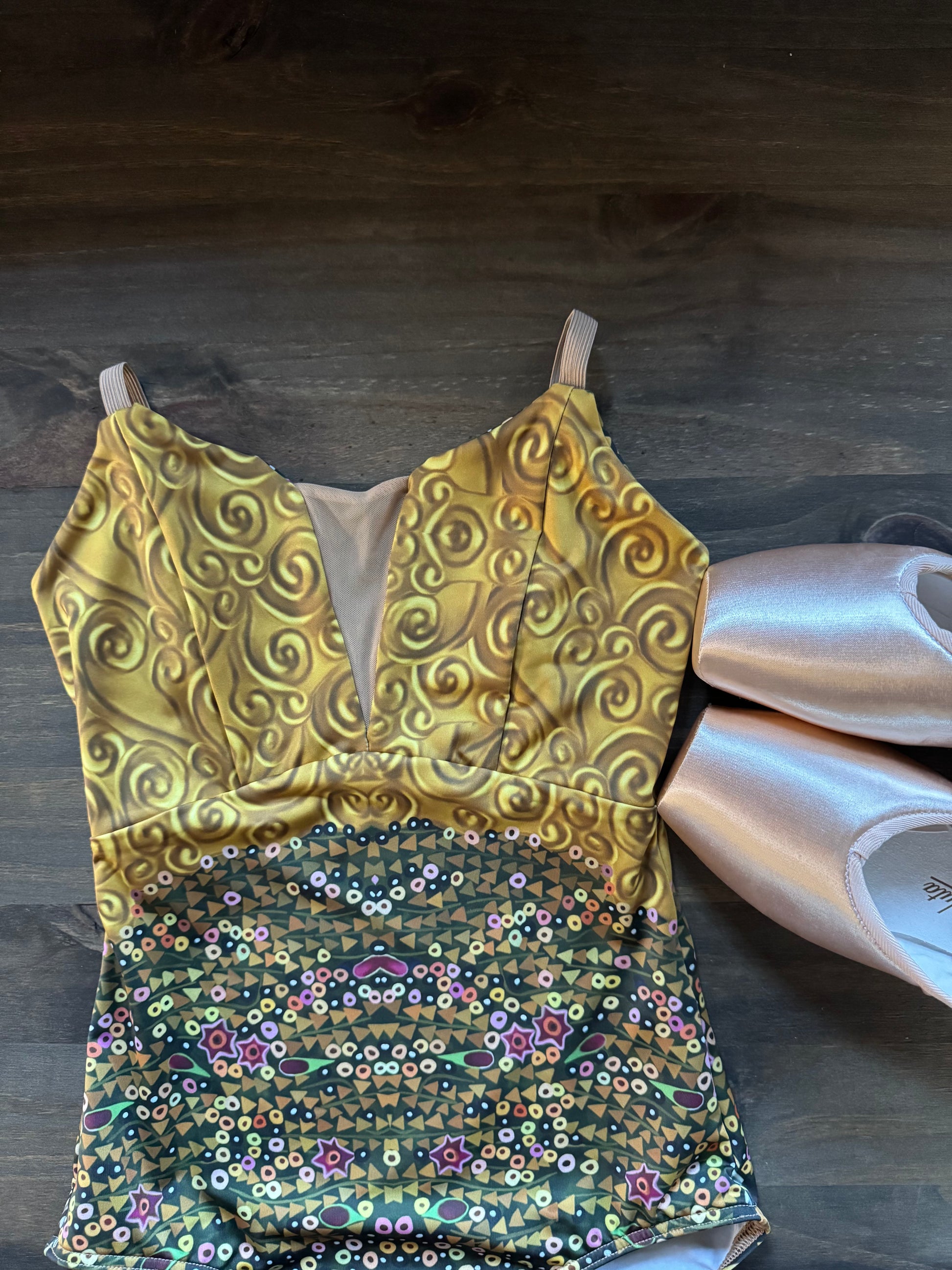 A sleeveless leotard or dance top laid flat on a dark wood floor, with a metallic gold upper section featuring swirling patterns and a multicolored sequined floral lower section. Two pale pink satin ballet slippers are placed beside it.